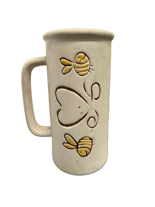 A beige ceramic mug with bee and heart designs on it, displayed on a flat surface.