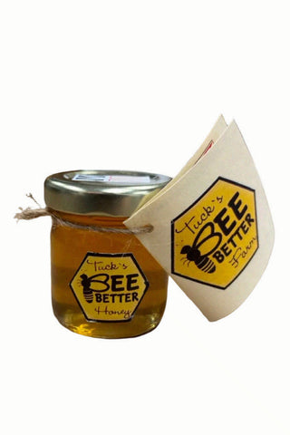 A small glass jar of honey sits on a wooden tabletop, with two larger jars blurred in the background. The front jar has a gold lid, a hexagonal yellow label reading 'Tucko's BEE Better Honey,' and a paper tag attached by twine.