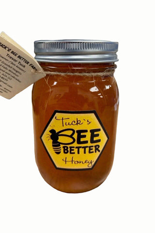 A glass mason jar filled with amber honey sits on a light wood table, featuring a hexagonal yellow label that reads 'Tuck's BEE BETTER Honey' with a bee graphic. A paper hang tag is attached to the side, and another honey bottle is blurred in the background.