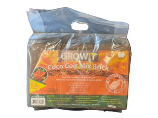 A packaged brick of Grow It Coco Coir Mix, held in front of a background that includes various objects. The packaging is primarily orange and white with text and branding.